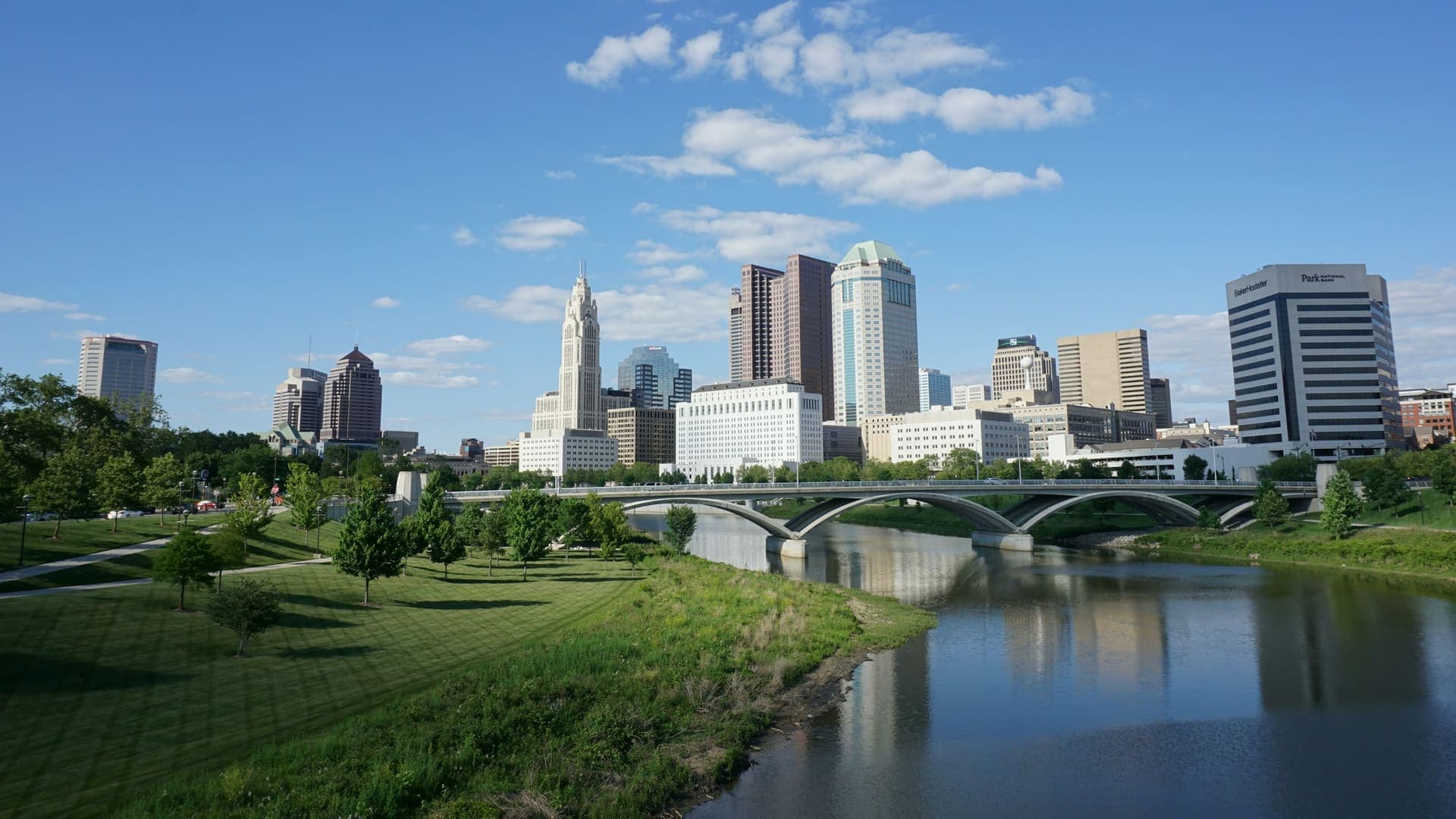 The downtown Columbus, Ohio skyline above the Scioto River and green park lawns on a sunny day.