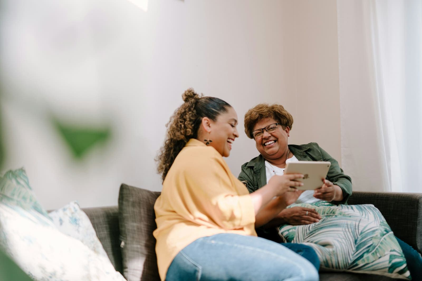 A younger woman and an older woman laugh together on a sofa while looking at a tablet.