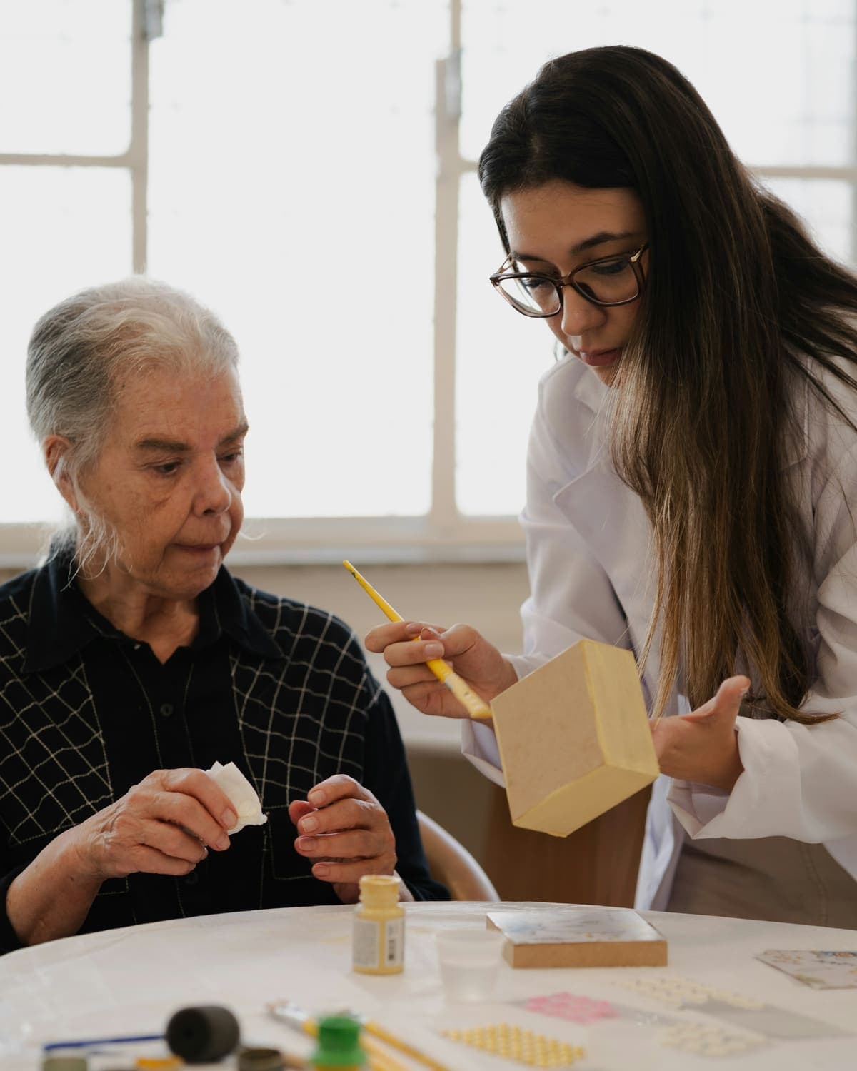 A young woman helps an older woman paint a small wooden box at a table by a bright window.